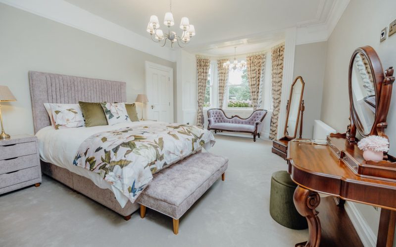 Elegant Cherry Room at Bettystown House featuring a plush bed with floral bedding, a classic wooden vanity with mirror, a soft velvet bench, and a bay window with a vintage sofa and garden view.